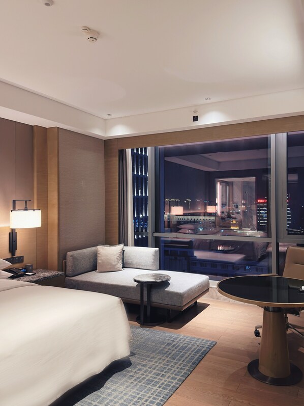 Habitación de hotel moderna con una cama grande, un sofá, una mesita y vistas a la ciudad iluminada de noche.