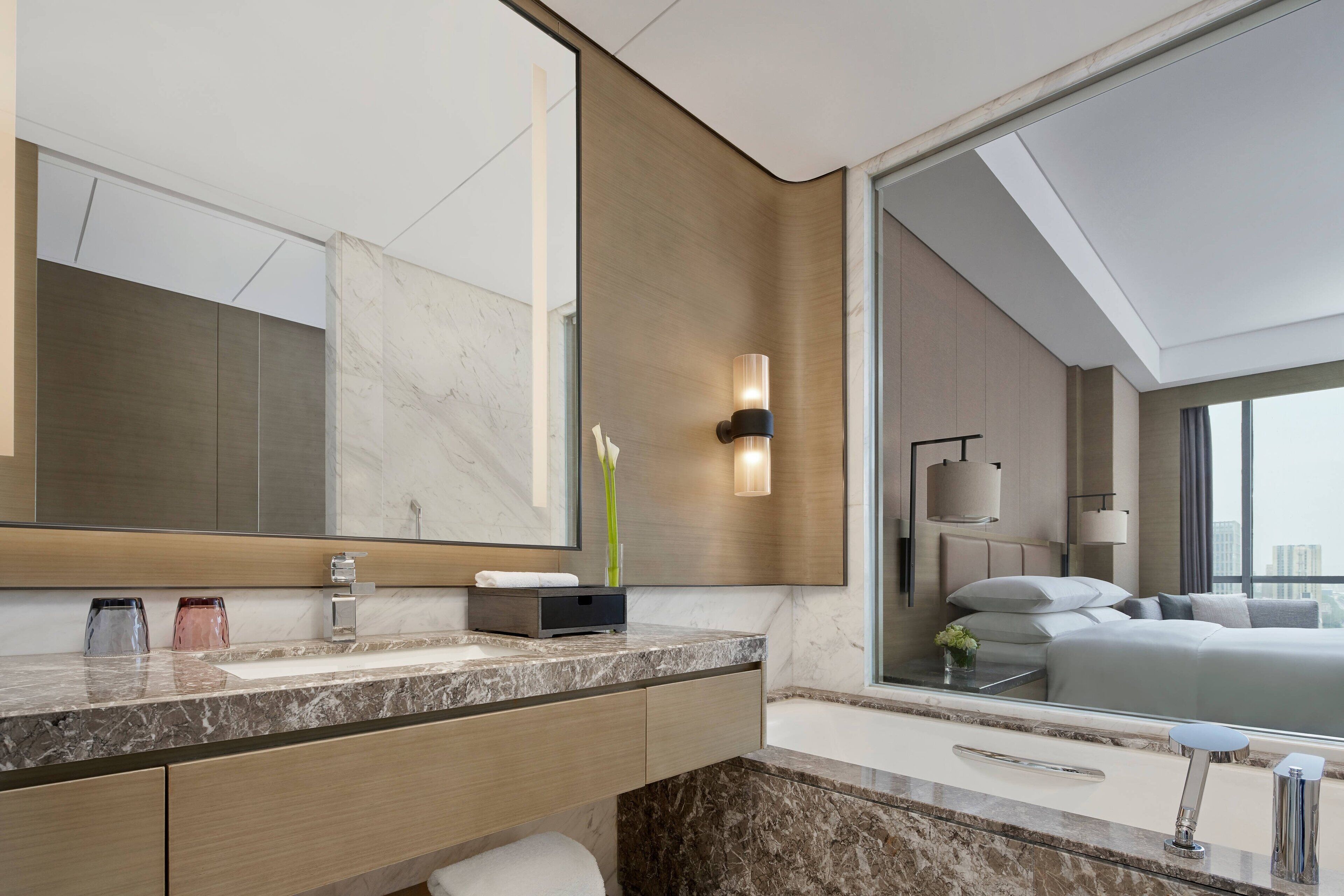A modern bathroom with a large mirror, marble countertop, and a view of a bedroom with a bed and window.