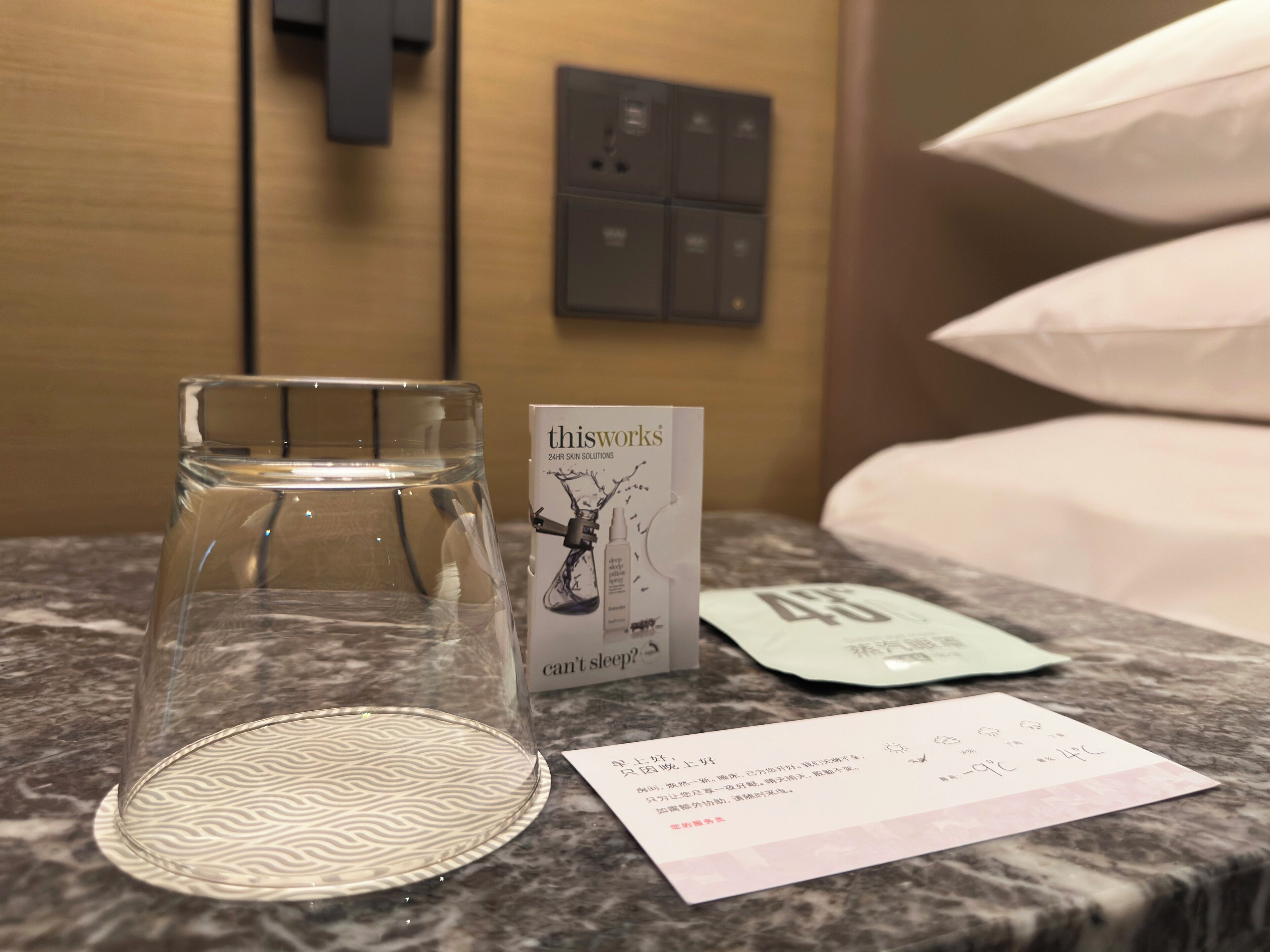 Ein Hotelzimmer mit einem Glas Wasser, einer Broschüre von 'thisworks' Skin Solutions, einem Temperaturanzeiger und einem Zettel auf dem Marmortisch.