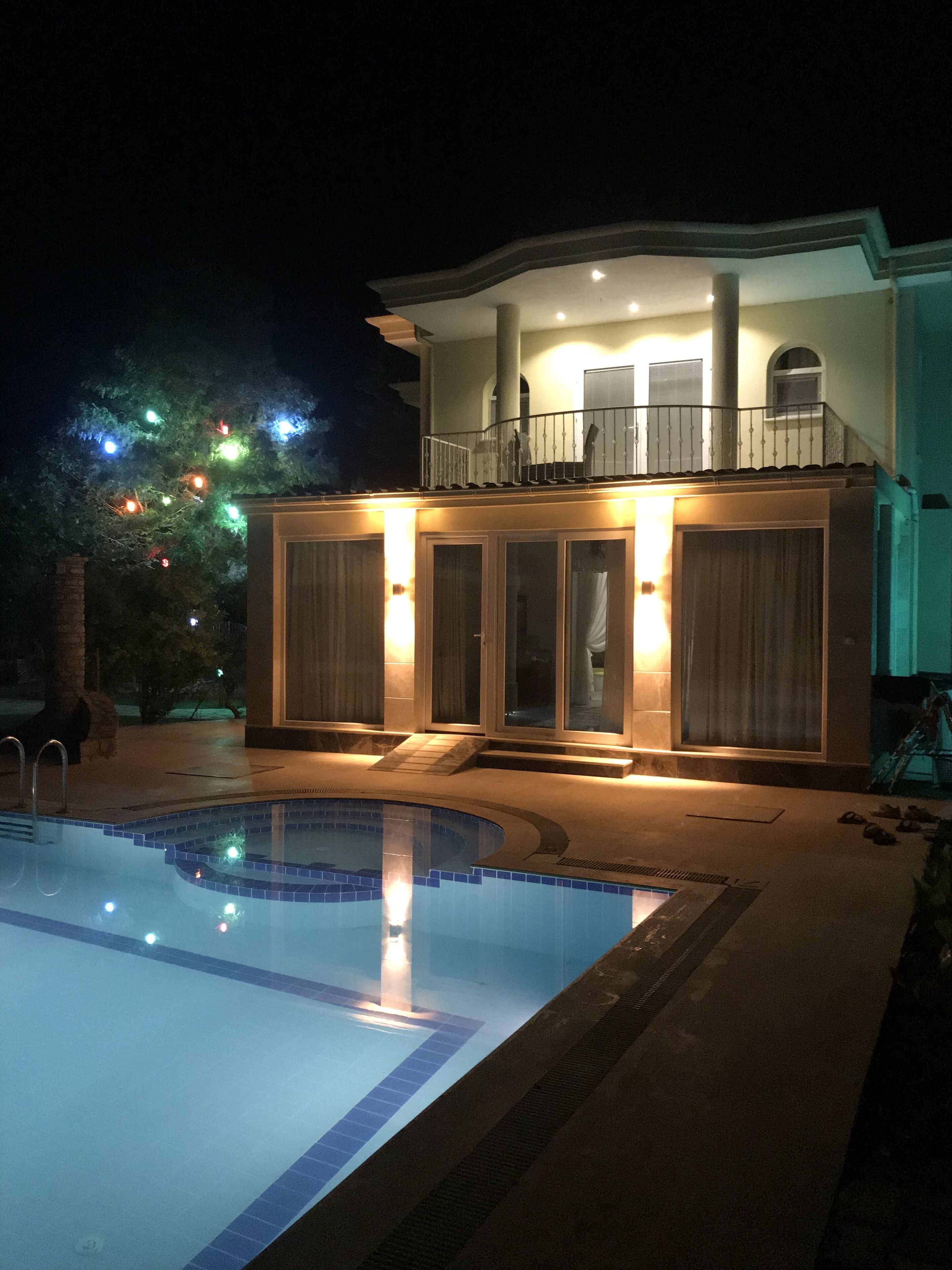 A poolside area with a swimming pool, a covered seating area with a balcony, and a tree with colorful lights.