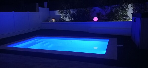 Une piscine rectangulaire dont l’intérieur est illuminé de lumière bleue, entourée d’un mur blanc et d’un petit jardin éclairé d’une lumière rose.