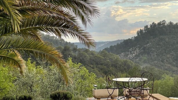 A patio with a table and chairs, surrounded by lush greenery and a view of a mountainous landscape.