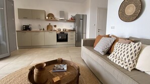 Una cocina moderna con horno empotrado, refrigerador y cafetera. Hay una zona de comedor con una mesa redonda de madera y un sofá con cojines estampados.
