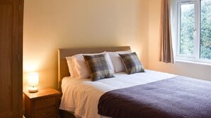 A neatly made bed with a white and brown bedspread, a wooden nightstand with a lamp, and a window with curtains. - Claife View - Parking, Balcony, 1 min walk to central Bowness (Bowness-on-Windermere)