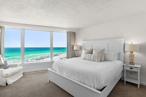 Habitación de hotel con un ventanal grande que ofrece vistas a la playa, una cama blanca, una mesita de noche con lámpara y una silla.