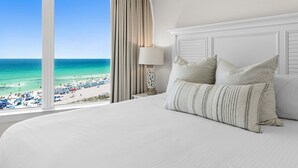 Habitación de hotel con una cama grande, una mesita de noche, una lámpara y vistas a la playa.