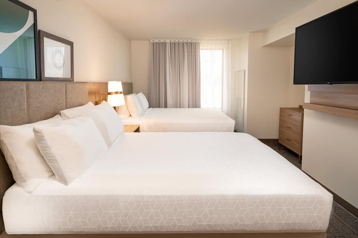 A hotel room with two beds, a TV, a desk, and a window with curtains.