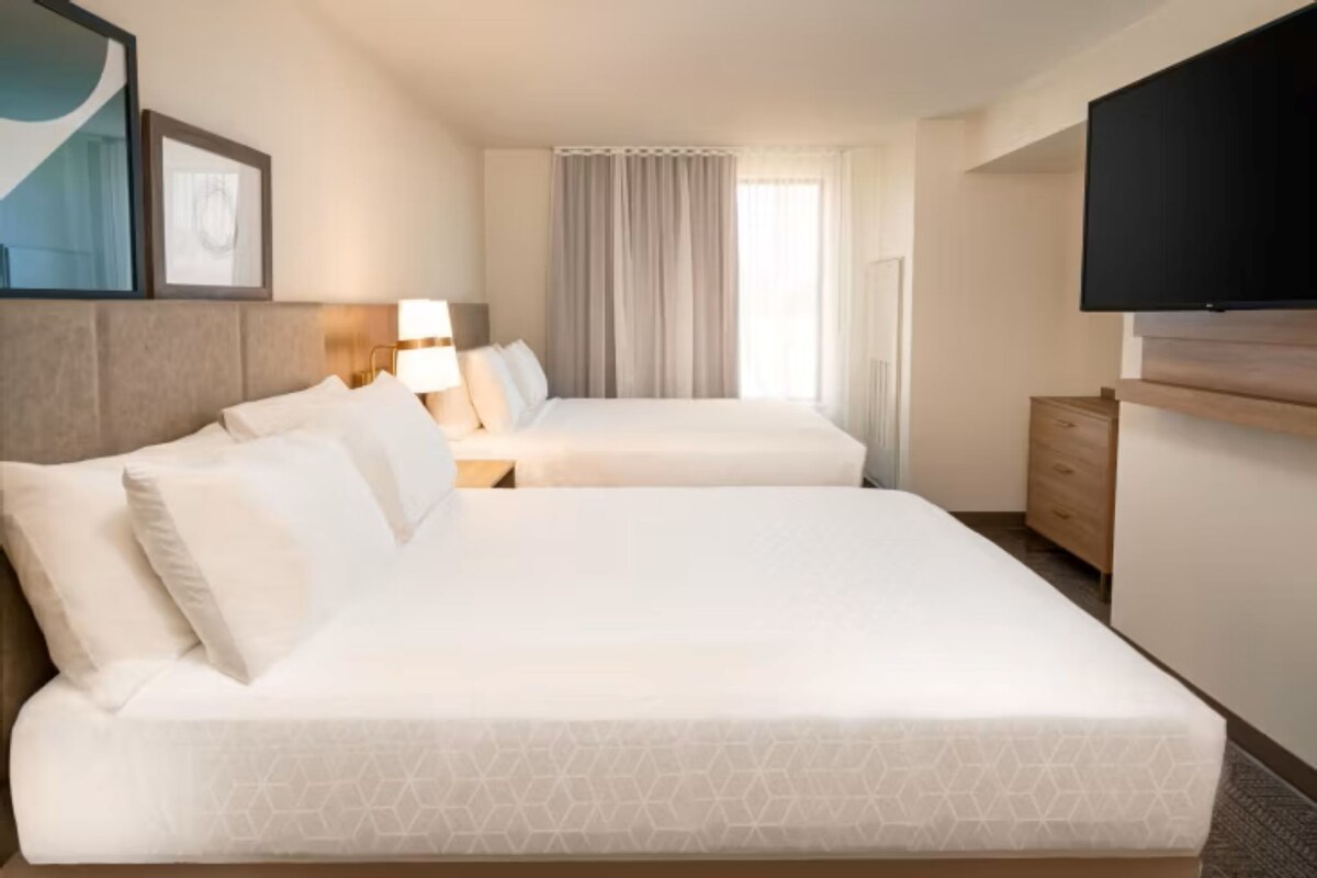 A hotel room with two beds, a TV, a desk, and a window with curtains.