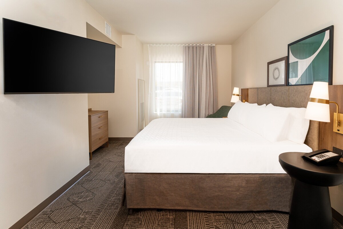 A hotel room with a large bed, a flat-screen TV mounted on the wall, a bedside table with a lamp, and a window with curtains.