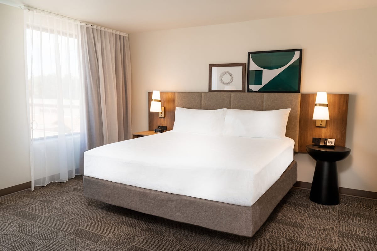 A hotel room with a large bed, two bedside tables with lamps, a headboard, and a window with sheer curtains.