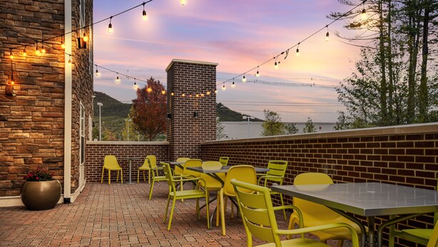 An outdoor patio with brick flooring, yellow chairs, and tables, featuring a brick wall and a view of a hill and water body. - Home2 Suites by Hilton North Conway (North Conway)
