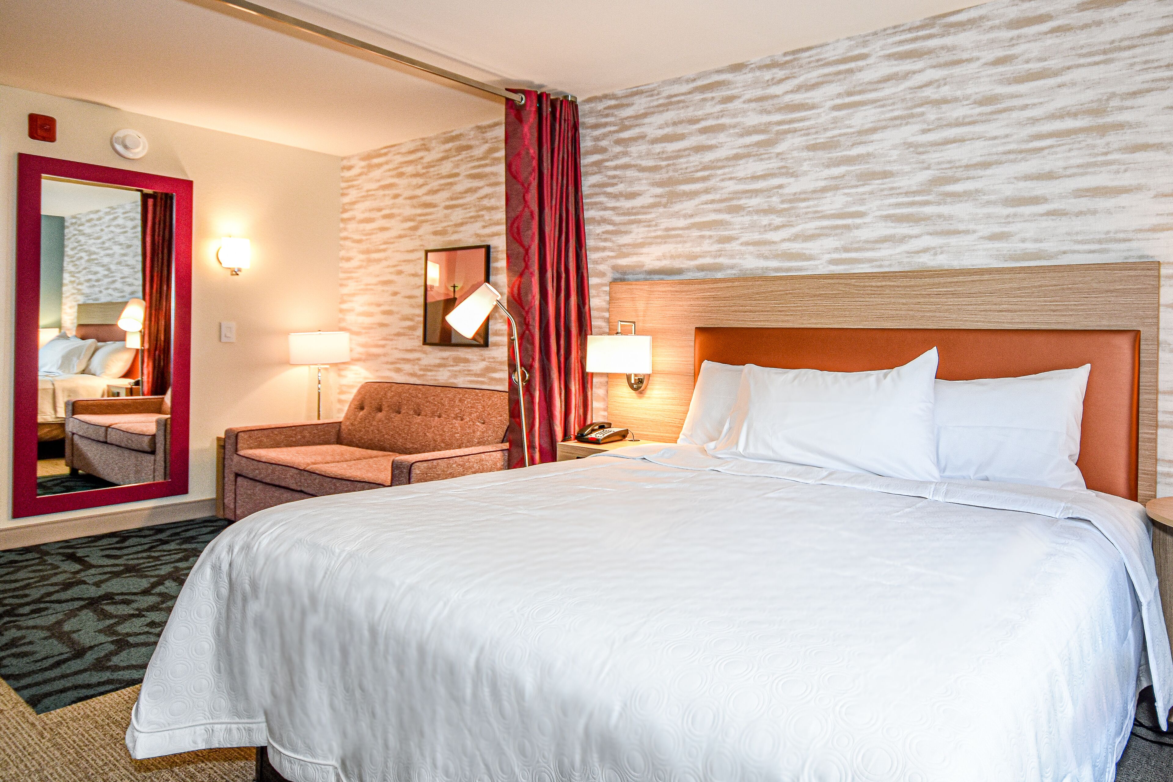 A hotel room with a large bed, a chair, a desk with a lamp, a telephone, and a mirror.