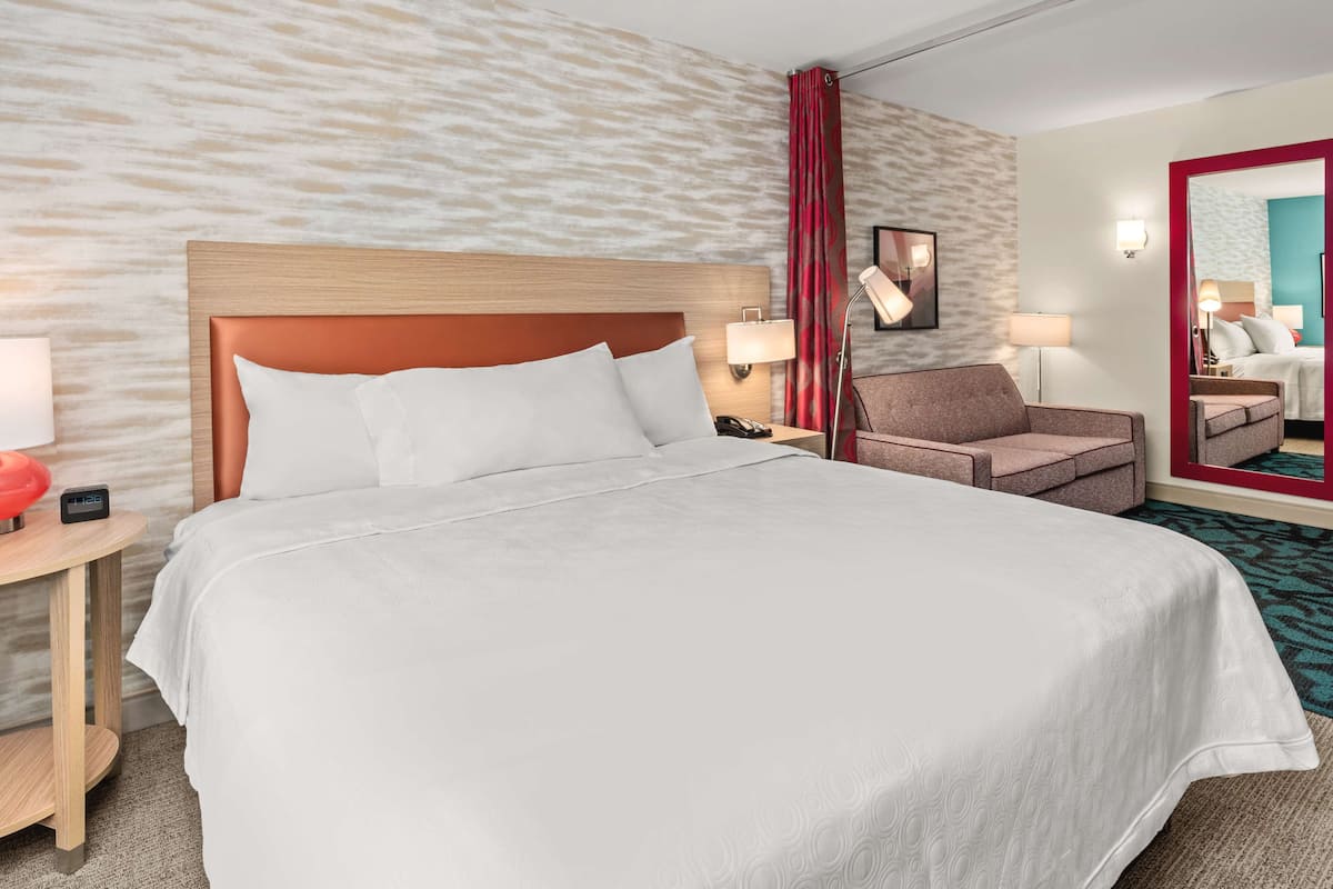 A hotel room with a large bed, a wooden headboard, a nightstand with a lamp, a chair, and a mirror.