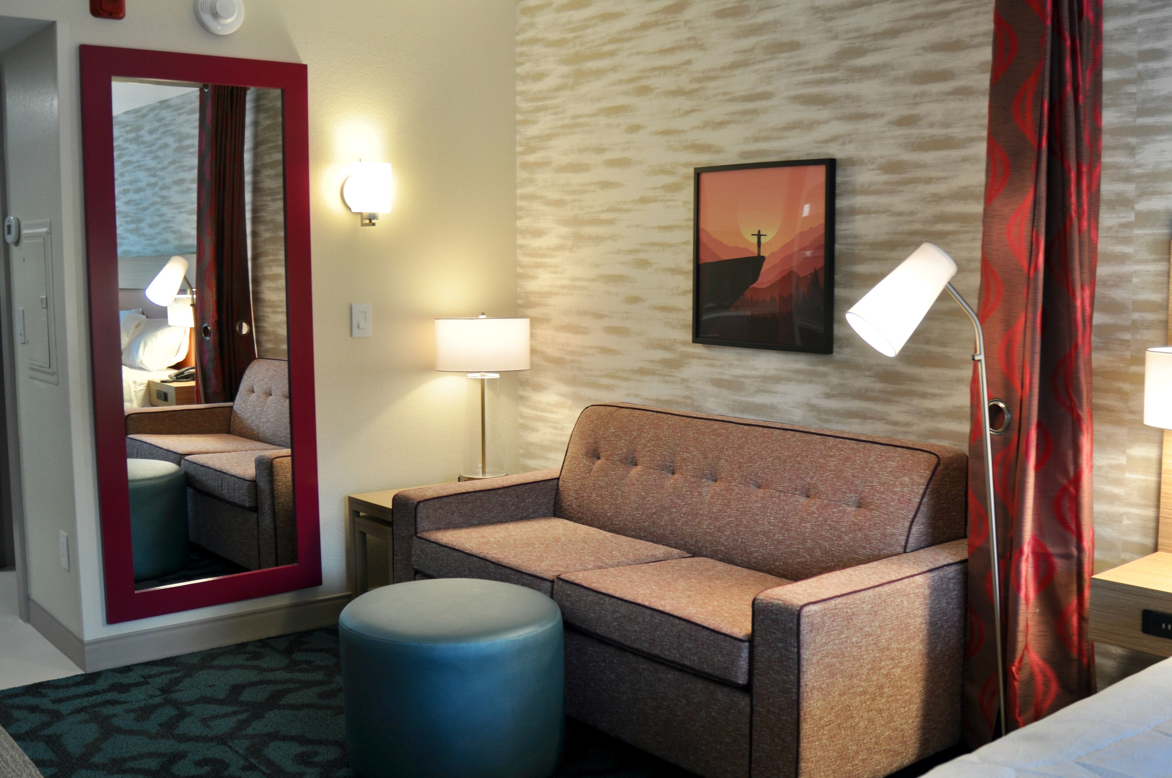 Una camera d'albergo con un divano, un poggiapiedi blu, uno specchio, una lampada e un quadro appeso al muro.