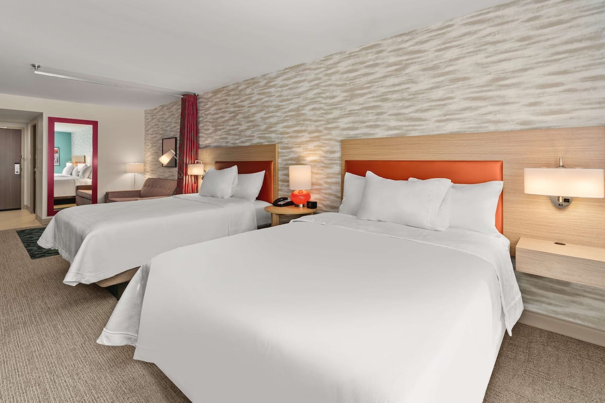 A hotel room with two beds, a wooden headboard, a nightstand with a lamp, and a red curtain.