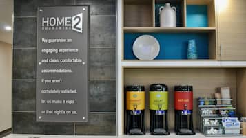 Zona de recepció d'hotel amb un cartell que indica una garantia de dues nits a casa, una estació de cafè amb tres dispensadors de begudes etiquetats com 'GASTRONÒMIC', 'DESCAFEÏNAT' i 'AIGUA CALENTA', i una estanteria amb objectes decoratius i subministraments.
