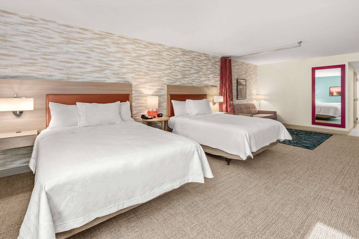 A hotel room with two beds, a wooden headboard, a red curtain, and a view of the city through a window.