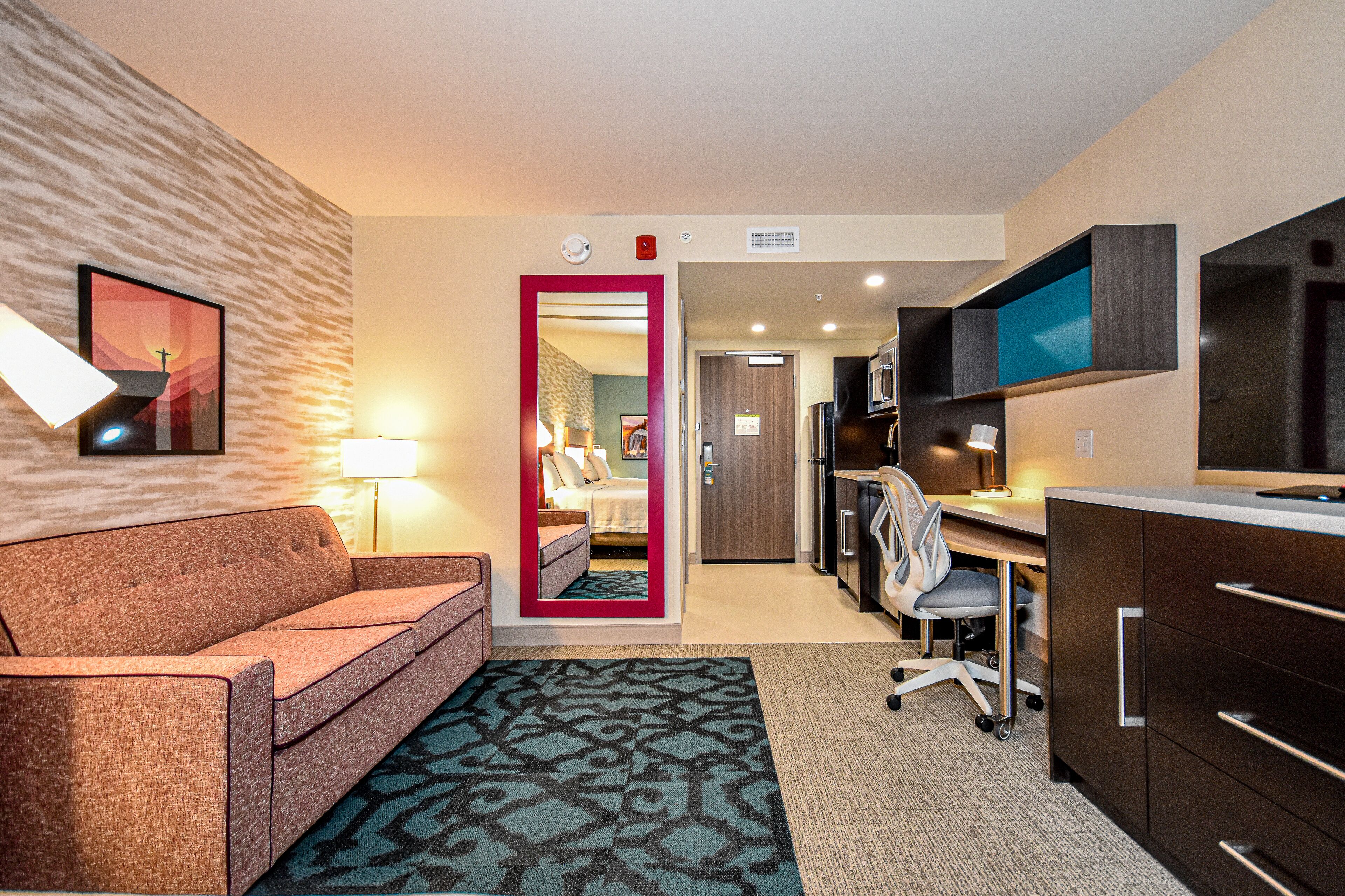 A hotel room with a sofa, a desk, a chair, a TV, and a kitchenette.