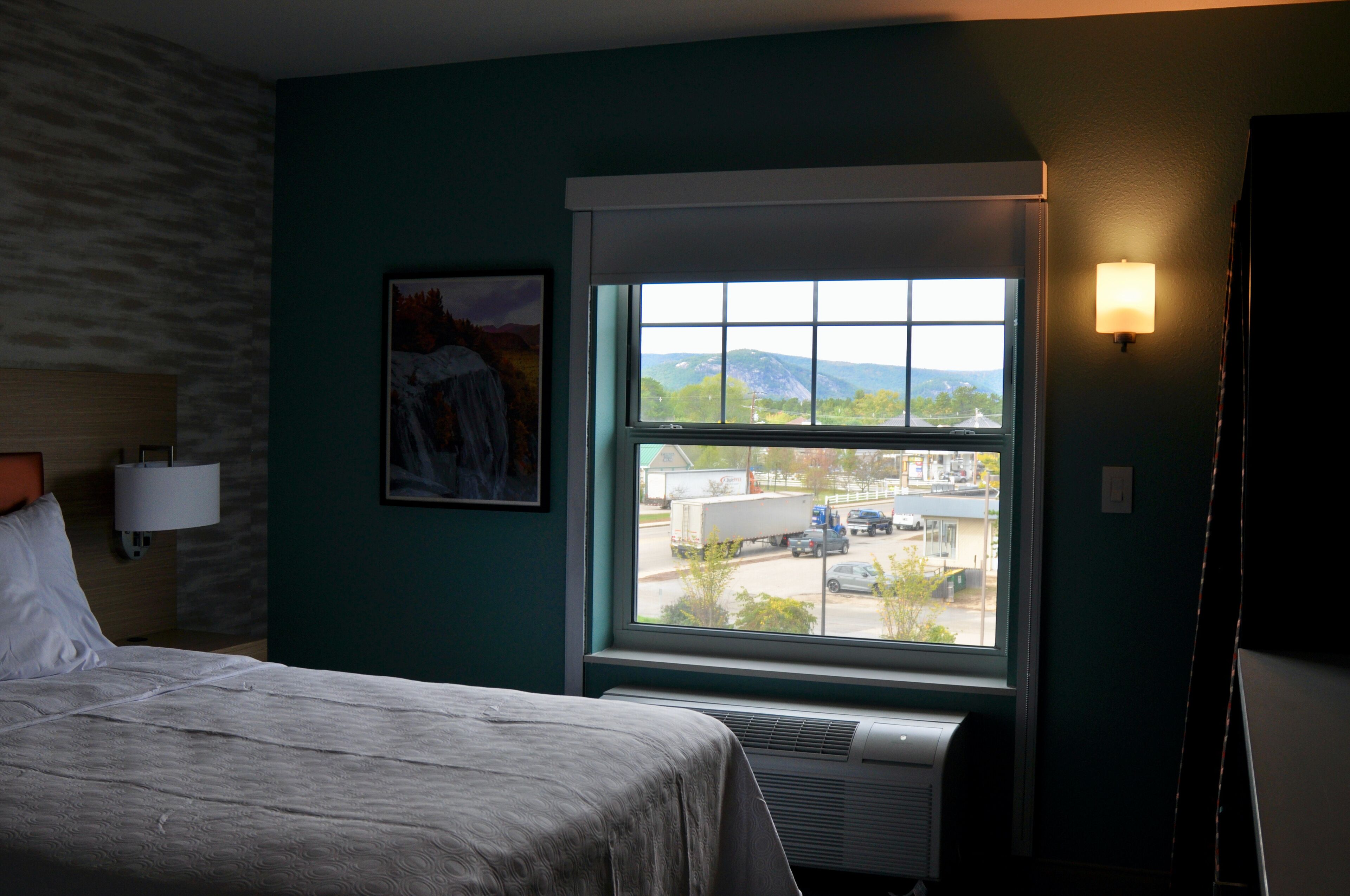 Quarto de hotel com cama, janela com vista para o exterior, luminária na parede e um quadro.