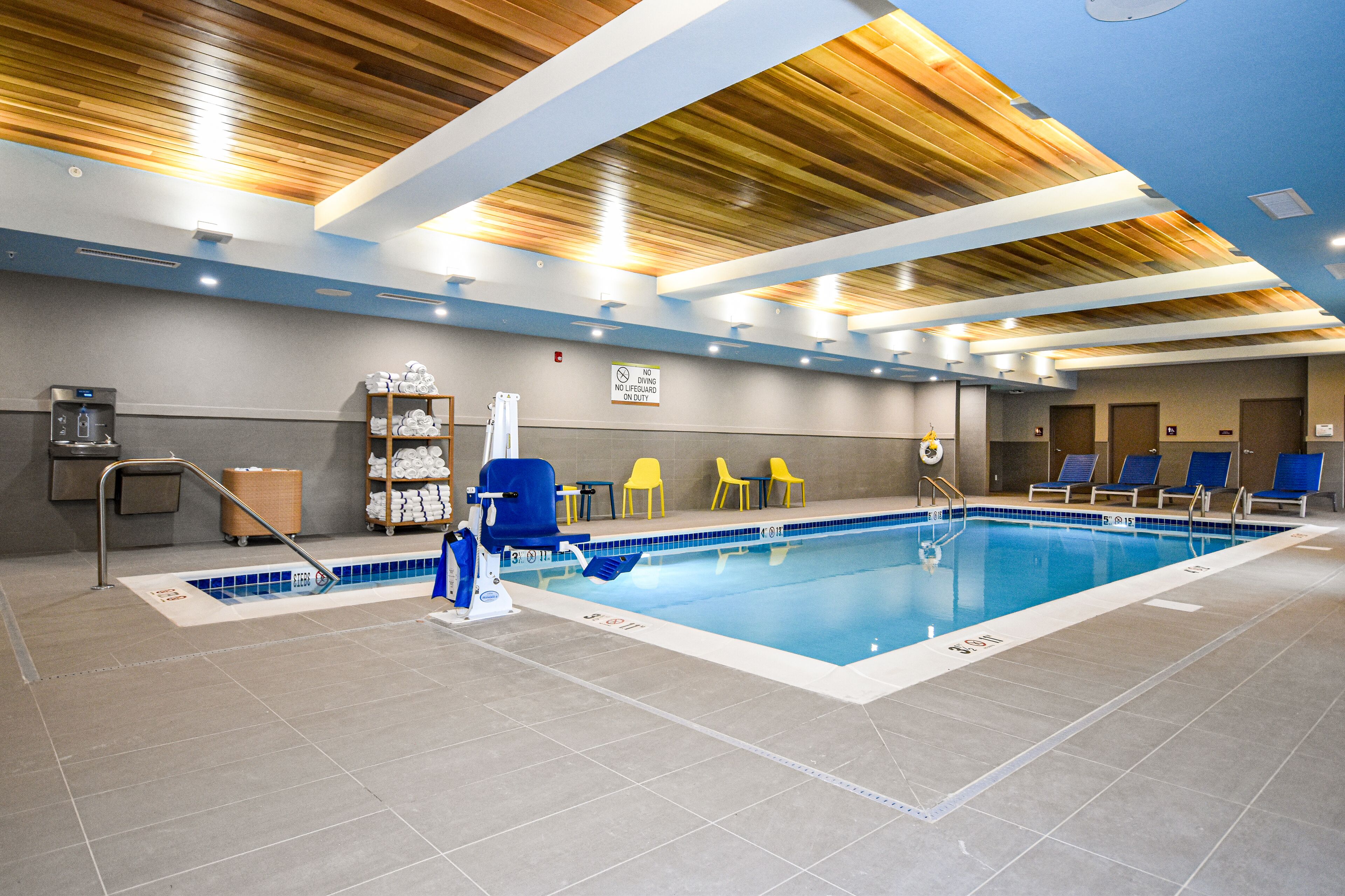 An indoor swimming pool with a wooden ceiling, blue and white tiled floor, and lounge chairs around the pool.