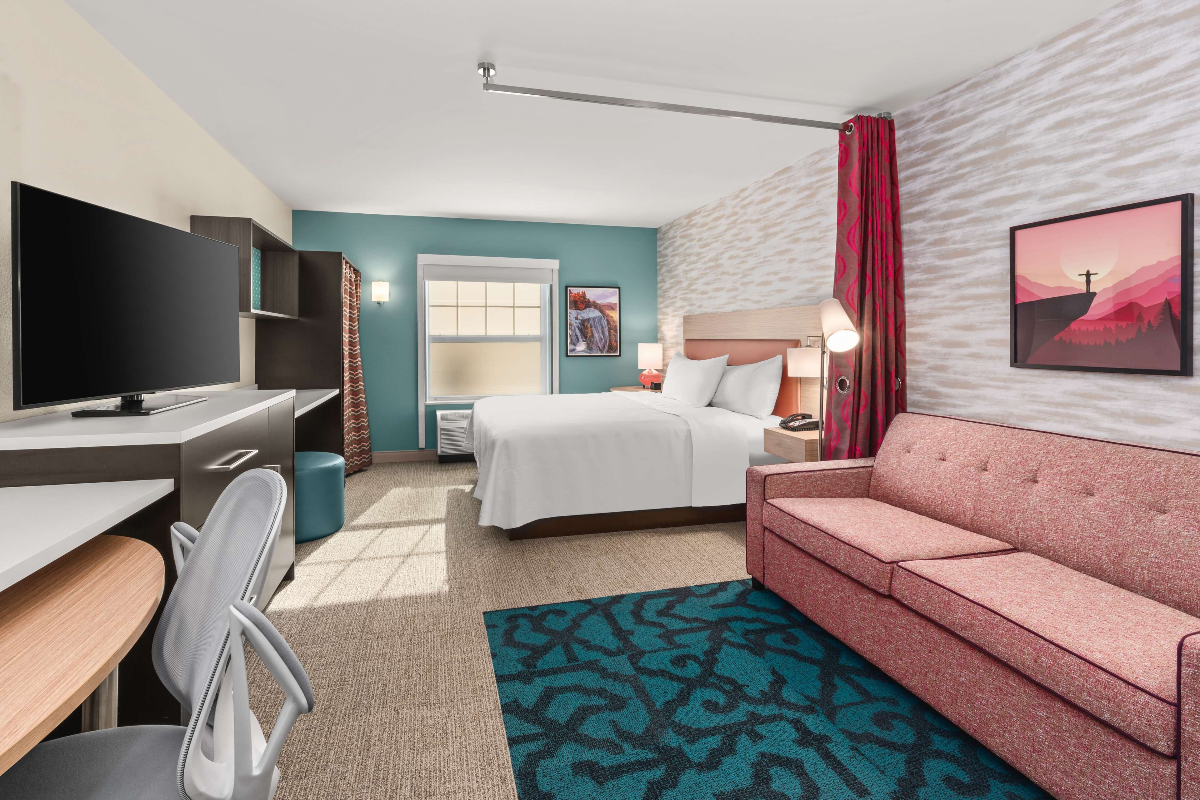 A hotel room with a bed, a desk with a chair, a television, a sofa, and a window with curtains.