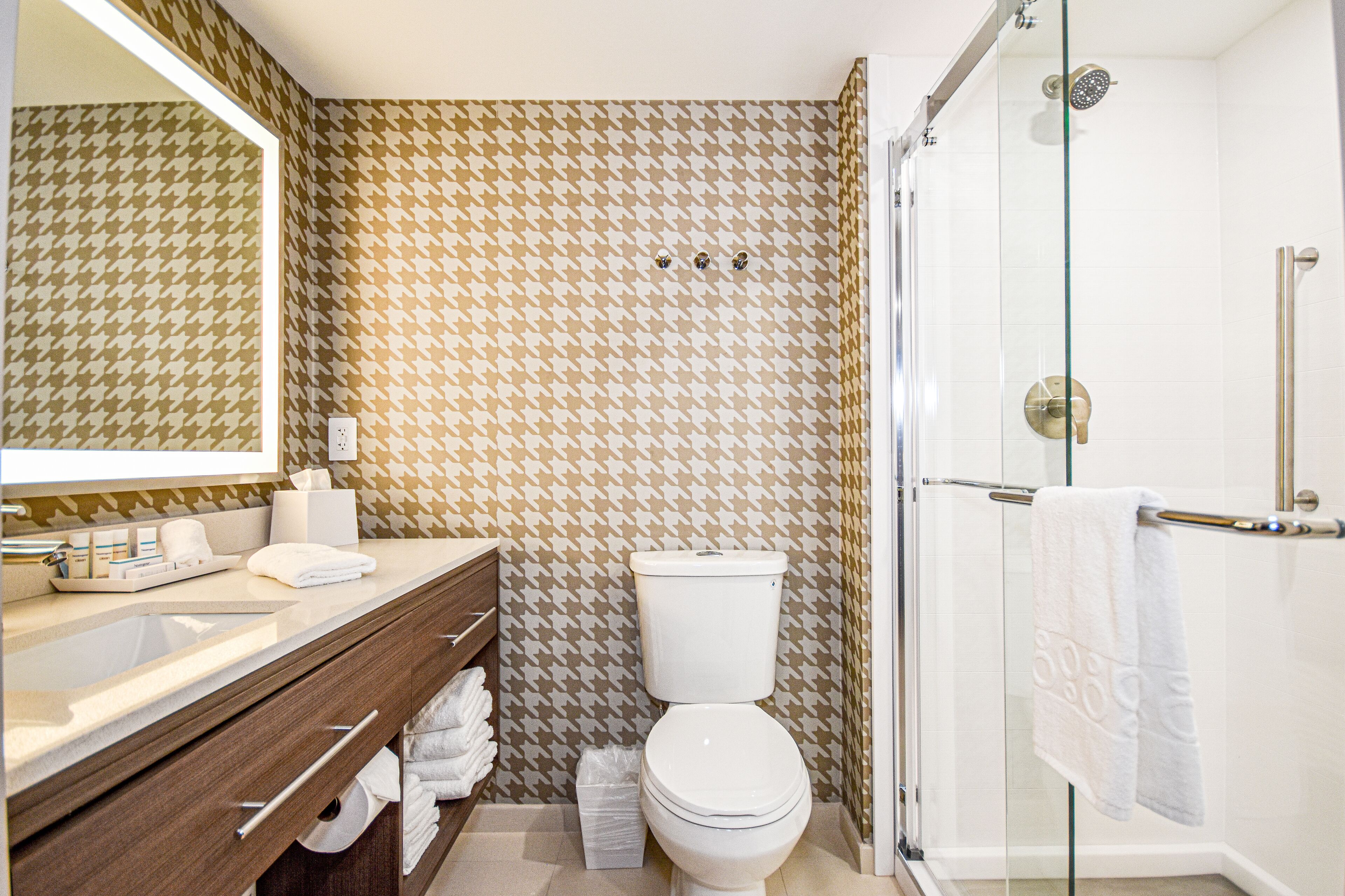 A bathroom with a houndstooth patterned wall, a white toilet, a glass-enclosed shower, and a wooden vanity with towels.