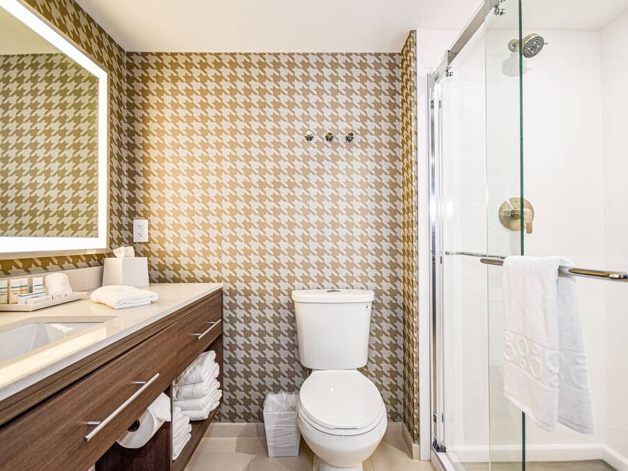 Une salle de bain avec un mur orné d’un motif à carreaux, des toilettes blanches, une douche en verre et un meuble-lavabo en bois avec des serviettes.