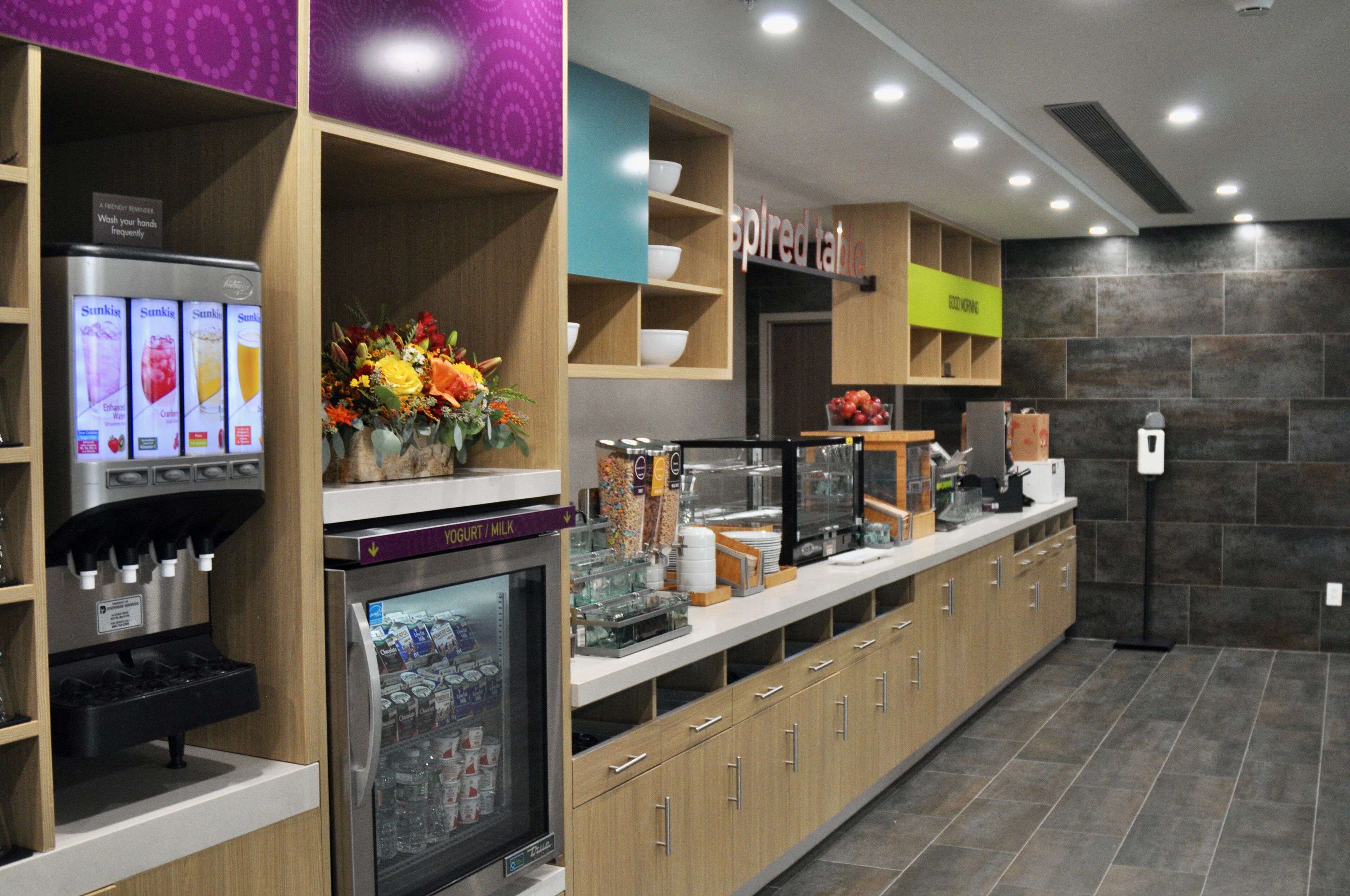 Una estación autoservicio para bebidas con un refrigerador, una máquina de café y diversas opciones de snacks.