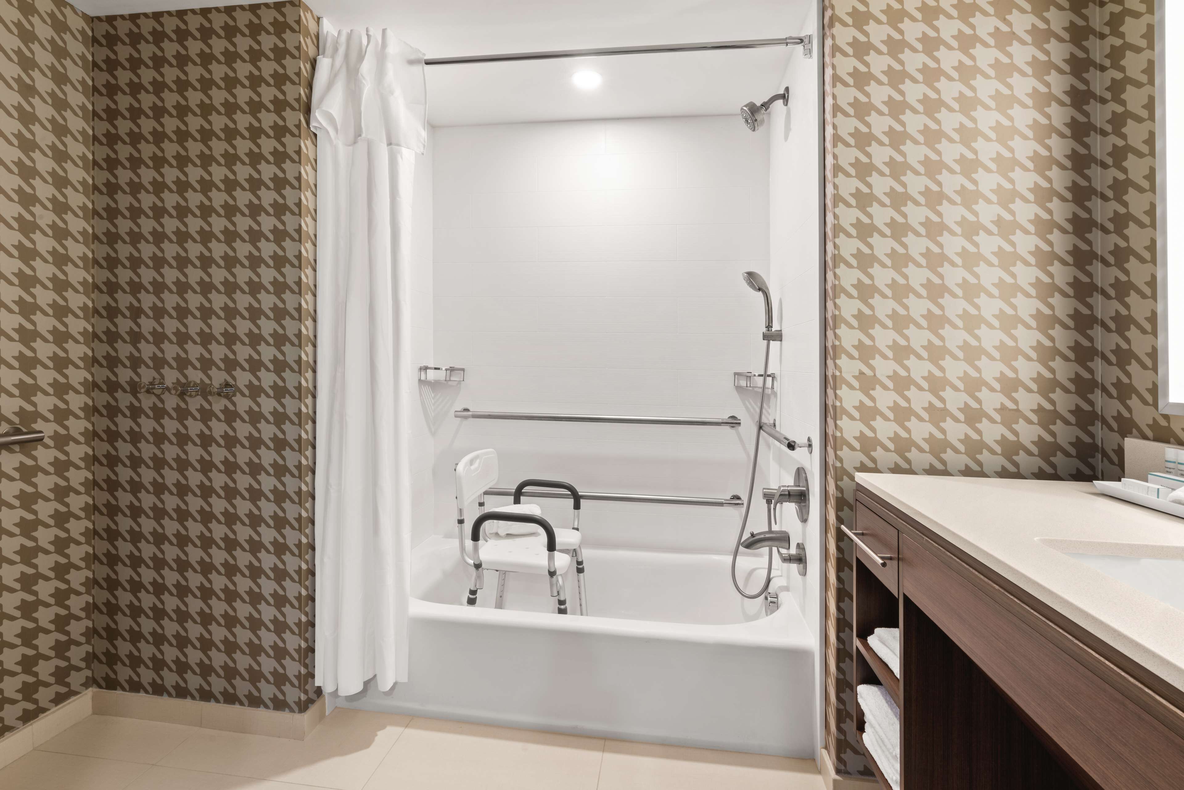 A modern bathroom with a walk-in shower, a white bathtub with a safety rail, and a vanity with a mirror.