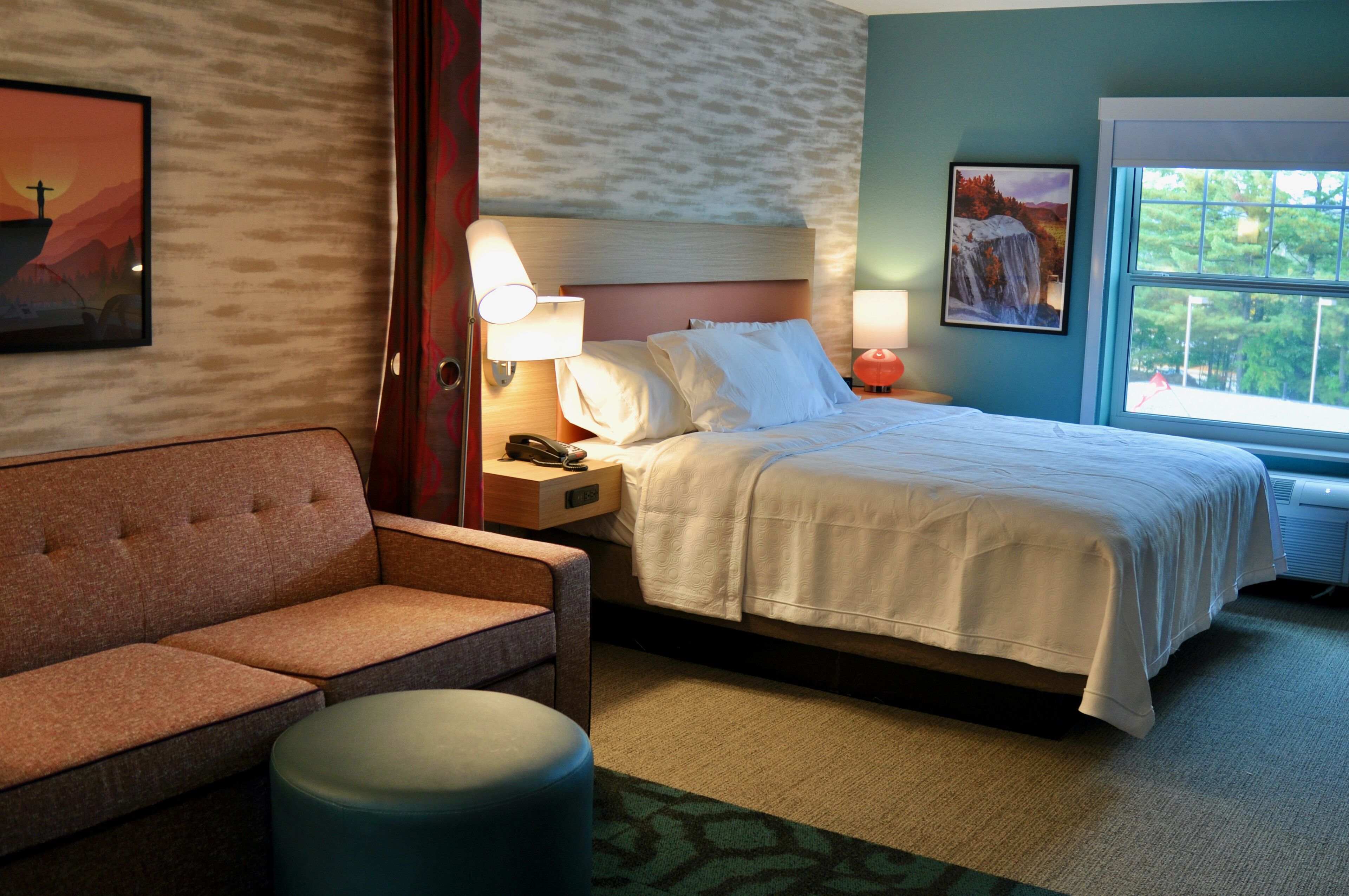 A hotel room with a bed, a sofa, a nightstand with a lamp, and a window with a view of trees.