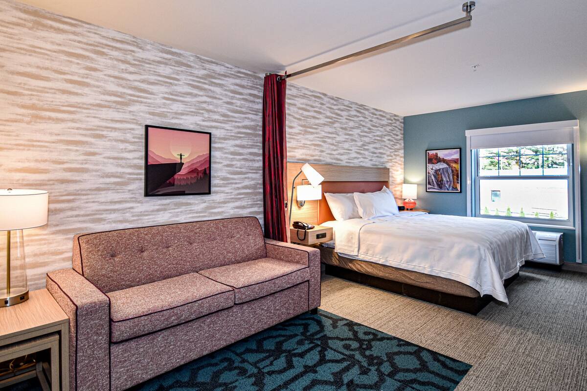 A hotel room with a bed, a sofa, a nightstand with a lamp, a wall-mounted TV, and a window with a view.