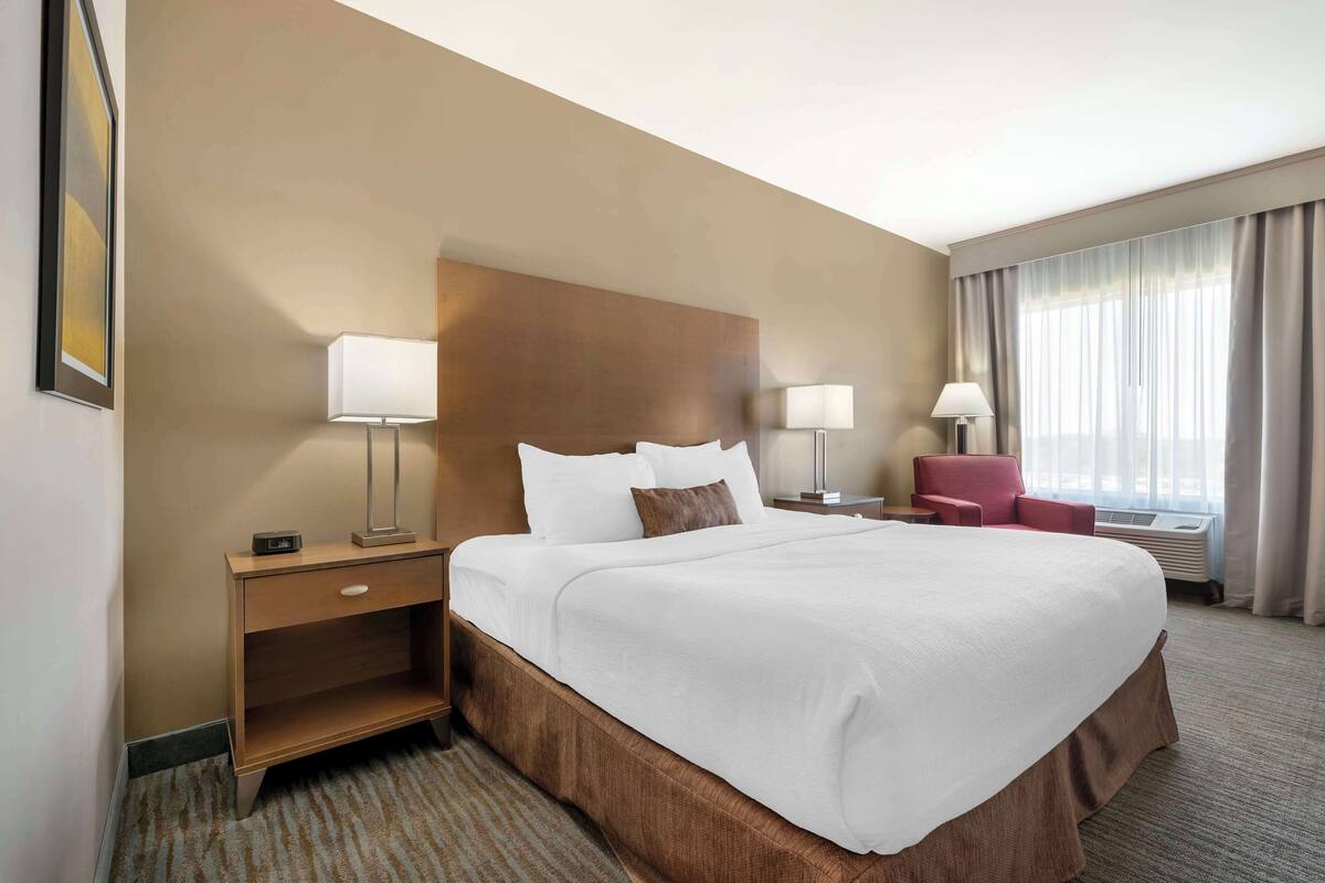 A hotel room with a large bed, two bedside tables with lamps, a red chair, and a window with curtains.