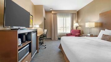 A hotel room with a large bed, a desk with a flat-screen TV, a chair, a lamp, and a window with curtains.