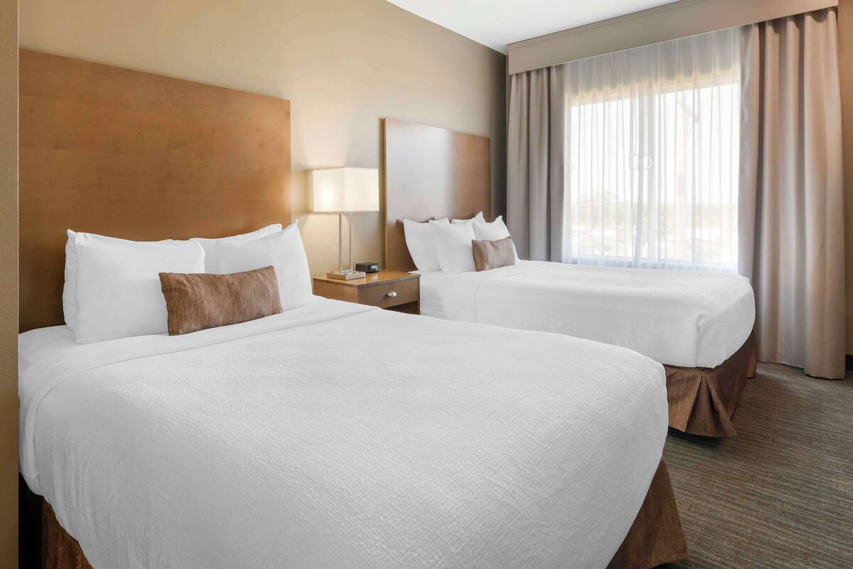 A hotel room with two large beds, a nightstand, a lamp, and a large window with curtains.