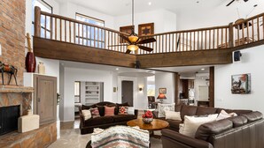 A spacious living area with a stone fireplace, a brown leather sofa, a wooden coffee table, and a high ceiling with exposed beams and a ceiling fan. - Happy Valley Hideaway | Resort-like home with amazing views & walking distance to hiking trails! (Phoenix)