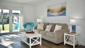 Una sala de estar con un sofá color beige, un sillón azul, mesitas blancas y un cuadro con una escena de playa.