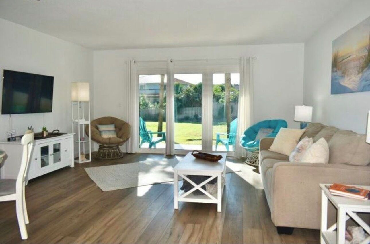 A spacious living room with a sofa, a coffee table, and a TV. There are two chairs and a lamp. The room has large windows with a view of greenery outside.