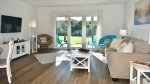 Amplia sala de estar con sofá, una mesa de centro y televisor. Hay dos sillones y una lámpara. La habitación tiene ventanas grandes con vistas a la vegetación exterior.