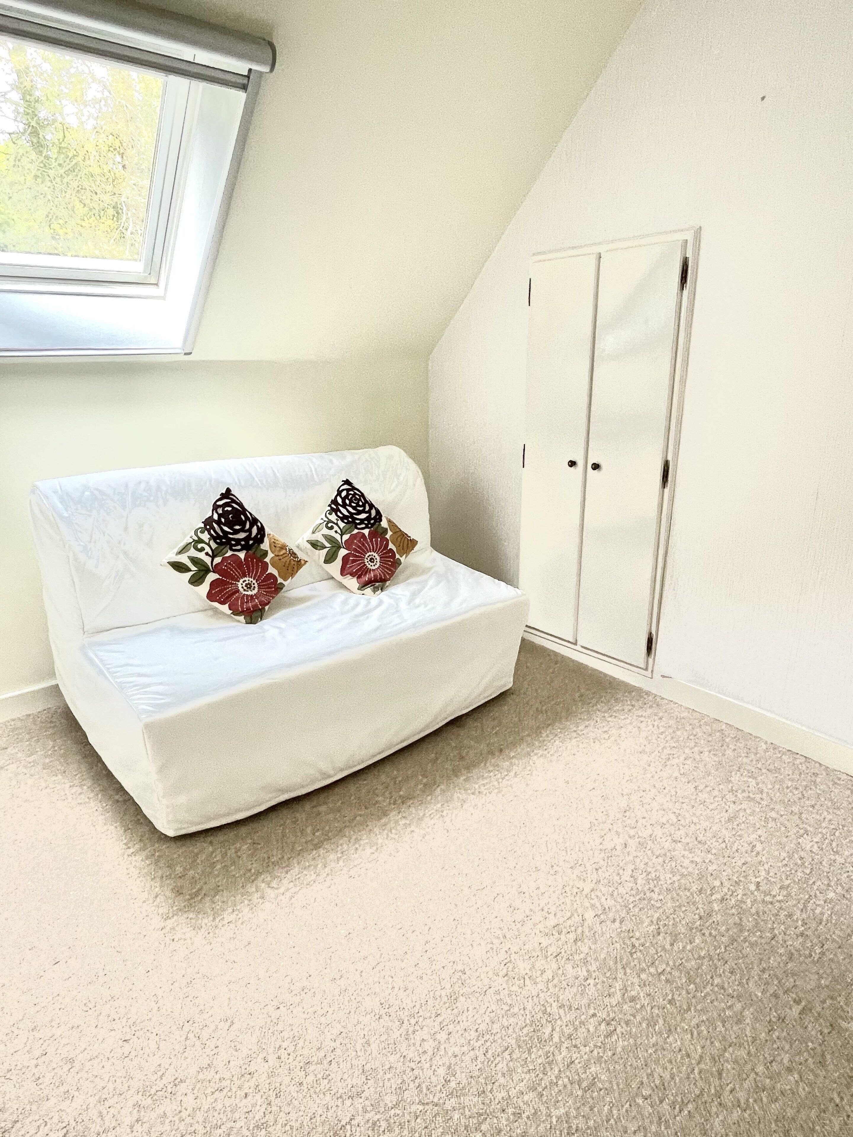 Une petite pièce blanche en forme de L, avec un canapé et deux coussins, une armoire et une fenêtre.