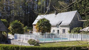 A stone house with a swimming pool, surrounded by a white fence, and a lush green landscape.