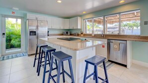 A modern kitchen with white cabinets, a stainless steel refrigerator, a microwave, and a sink. There are three bar stools and a large window with a view of palm trees.