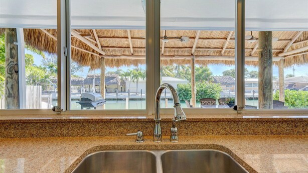 A kitchen with a granite countertop, stainless steel sink, and a view of a pool area with thatched roofs.