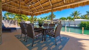 A covered outdoor dining area with a thatched roof, surrounded by palm trees and a pool.