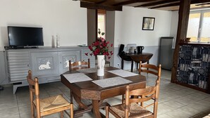 A dining area with a wooden table, four chairs, a vase with flowers, a television on a cabinet, and a refrigerator. - Rent Sarlat in Perigord  (Sarlat-la-Canéda)