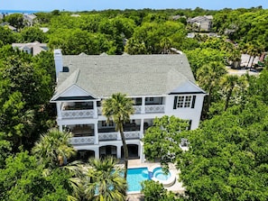 A large, two-story house with a swimming pool, surrounded by lush greenery and palm trees.