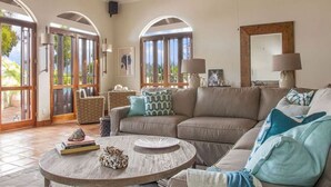 Ein geräumiges Wohnzimmer mit einem beige farbenen Sofa, einem runden Couchtisch und einem gemusterten Teppich. An beiden Seiten des Sofas befinden sich zwei Lampen, und an der Wand hängt ein großer Spiegel.