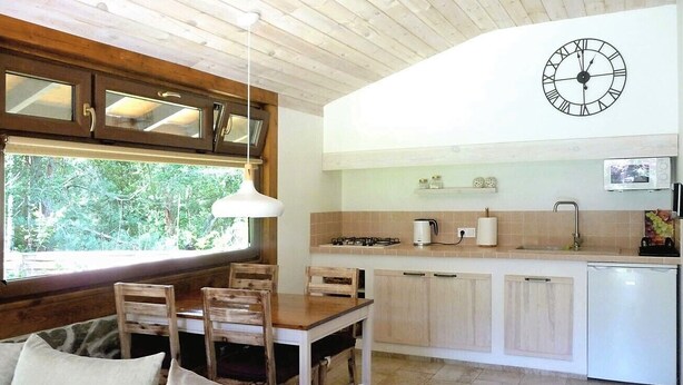 Une cuisine chaleureuse avec des meubles en bois, une table à manger avec des chaises, une horloge et une fenêtre donnant sur un espace verdoyant.