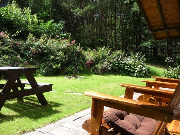 A wooden picnic table and chairs in a lush garden with dense foliage and trees in the background.