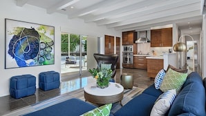 A modern living room with a sofa, ottomans, and a coffee table. There is a kitchen in the background with wooden cabinets and stainless steel appliances. - An Extra Hour of Sunshine, Southern California Desert (Rancho Mirage)