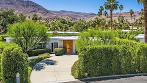 A modern building with a wooden facade, surrounded by lush greenery and palm trees, with mountains in the background. - An Extra Hour of Sunshine, Southern California Desert (Rancho Mirage)